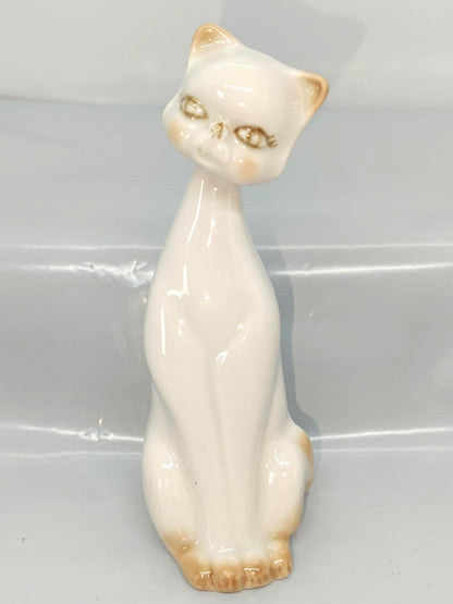 May include: A ceramic cat ornament in a seated position. The cat is mainly off-white with tan detailing on the ears, paws, and cheeks. The cat's head is slightly tilted, and it has detailed facial features, including eyes and a nose.